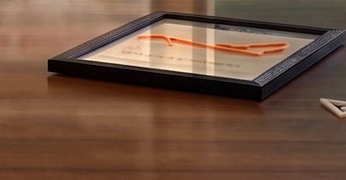 A race route 3D-printed in orange filament, mounted with a printed name inside a black shadow-box frame, lying on a wooden desk.